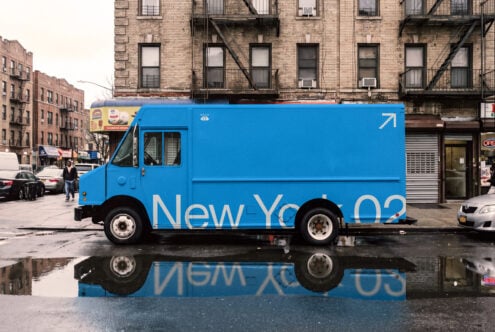 Bright blue delivery truck parked on NYC street with brick buildings reflected in puddle