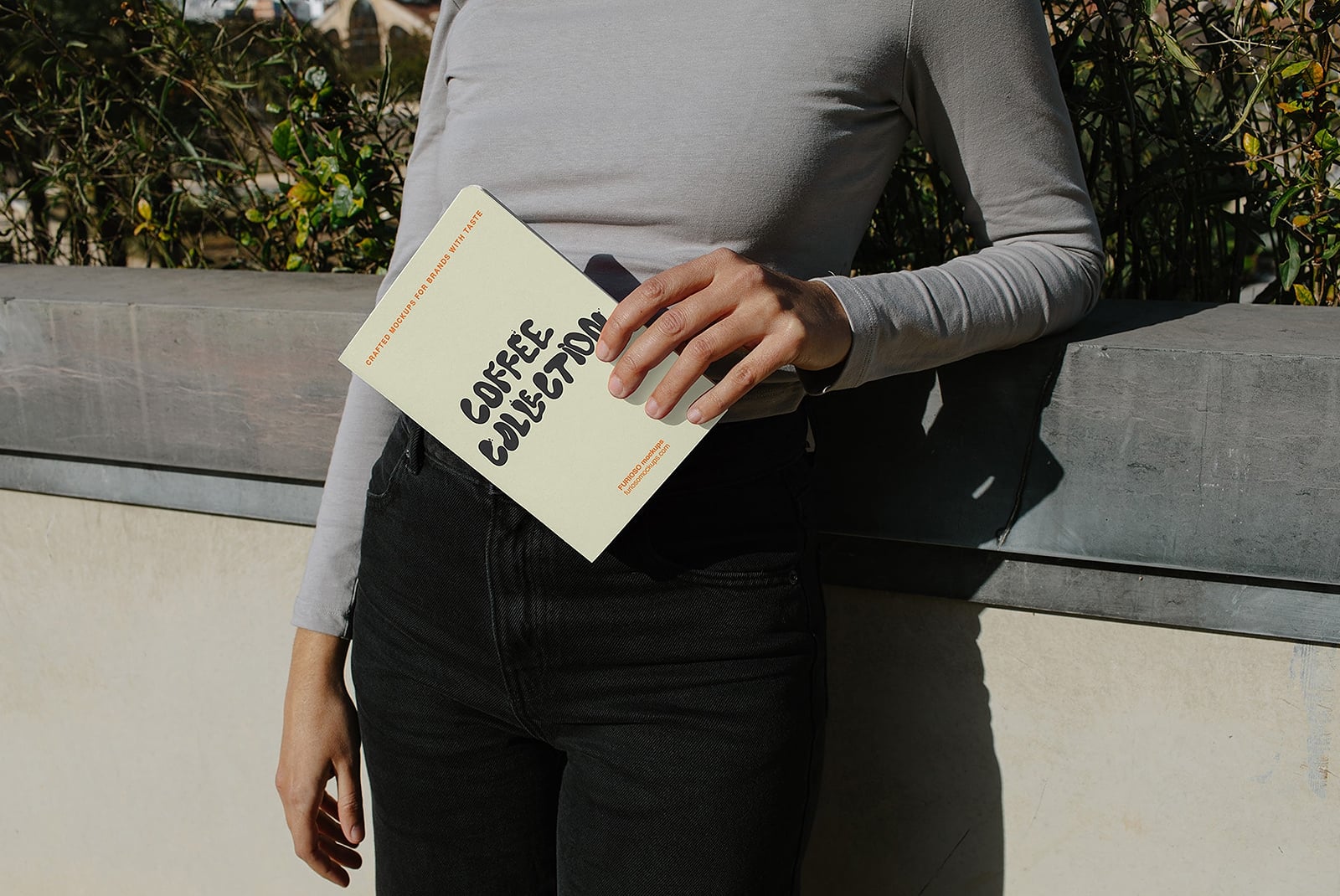 Person holding cream colored booklet with bold black typography in outdoor setting with plants