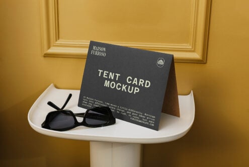 Tent card mockup on white pedestal table with black sunglasses, gold wall backdrop
