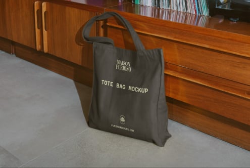 Black tote bag with logo text sitting on concrete floor against wooden cabinet with books