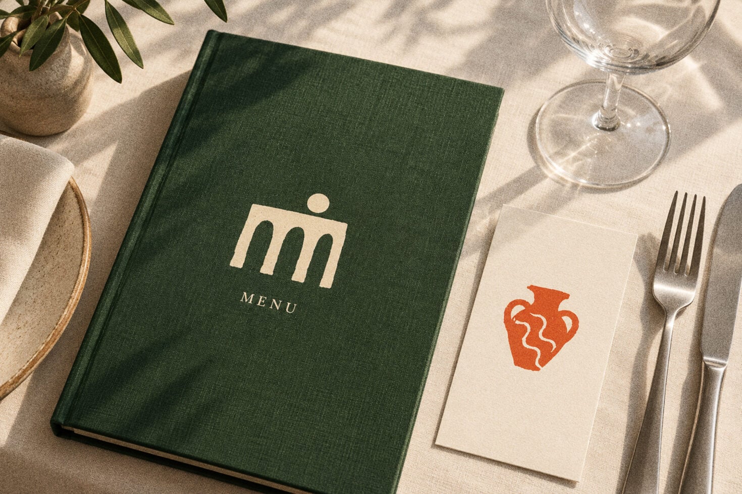 Green linen menu cover with minimalist arch logo, styled flat lay with wine glass, place setting