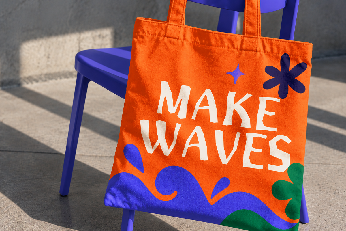 Bright orange tote bag with "Make Waves" text, blue waves, star, flower shapes next to blue chair