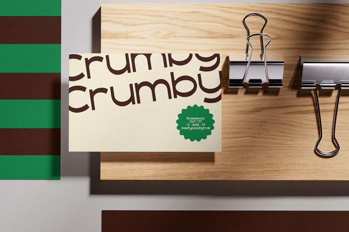 Cream notecard with "crumbly crumbly" text in brown, green badge logo, sitting on wood desk next to binder clips