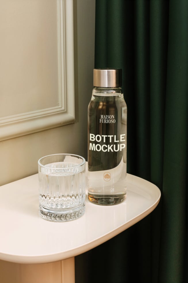 Dark glass bottle with gold cap next to crystal tumbler on round table, green curtain backdrop
