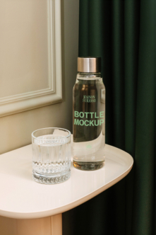 Dark glass bottle with gold cap next to crystal glass on round table, green curtain backdrop
