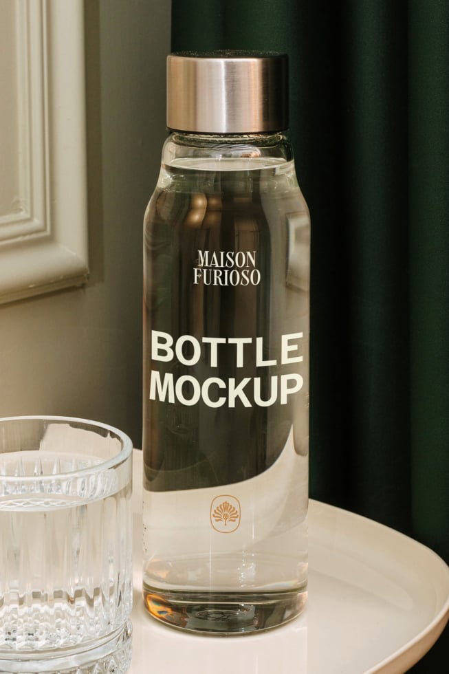 Glass bottle mockup with metallic cap, styled with crystal glass on neutral surface against green curtain backdrop