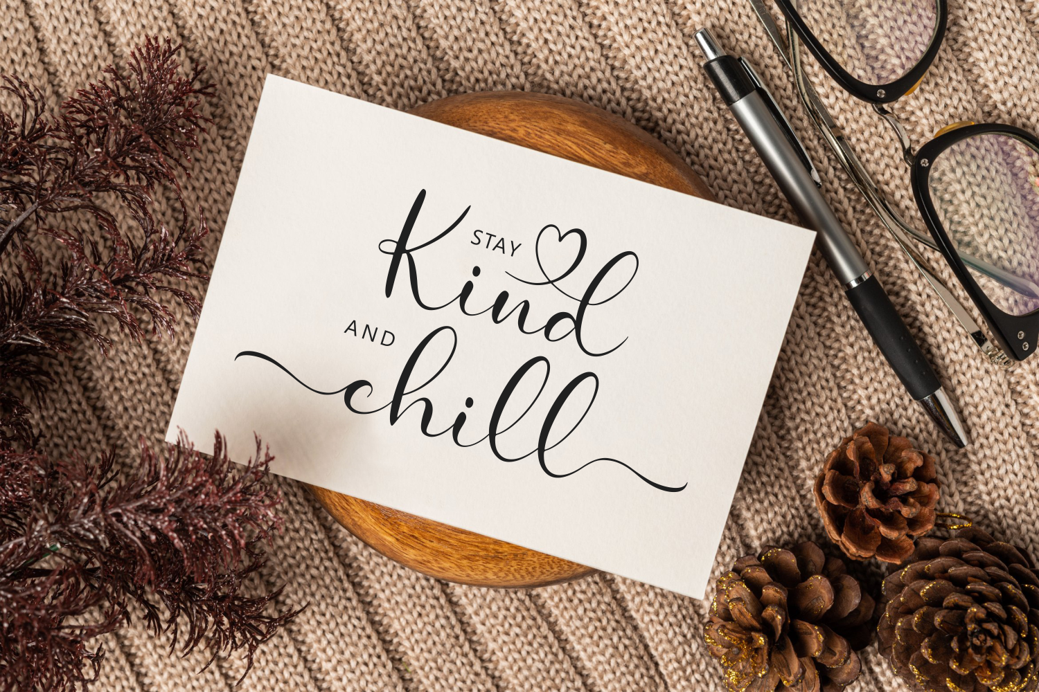 Card mockup with handwritten script saying stay kind, chill on beige linen with pen, glasses, dried flowers