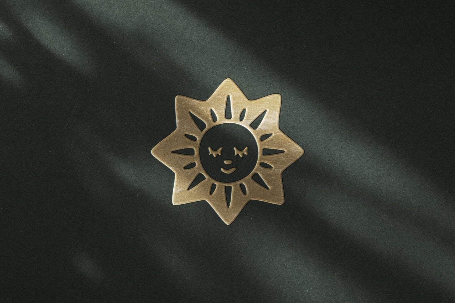 Brass sun face badge with closed eyes on textured dark green background with shadow overlay