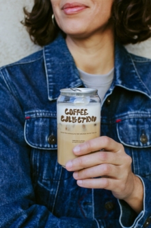 Woman in denim jacket holding iced coffee can mockup with brown lettering