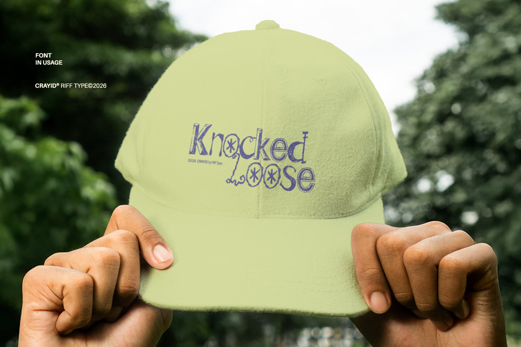 Lime green cap with "Knocked Loose" embroidered in purple, held up outdoors with blurred foliage background