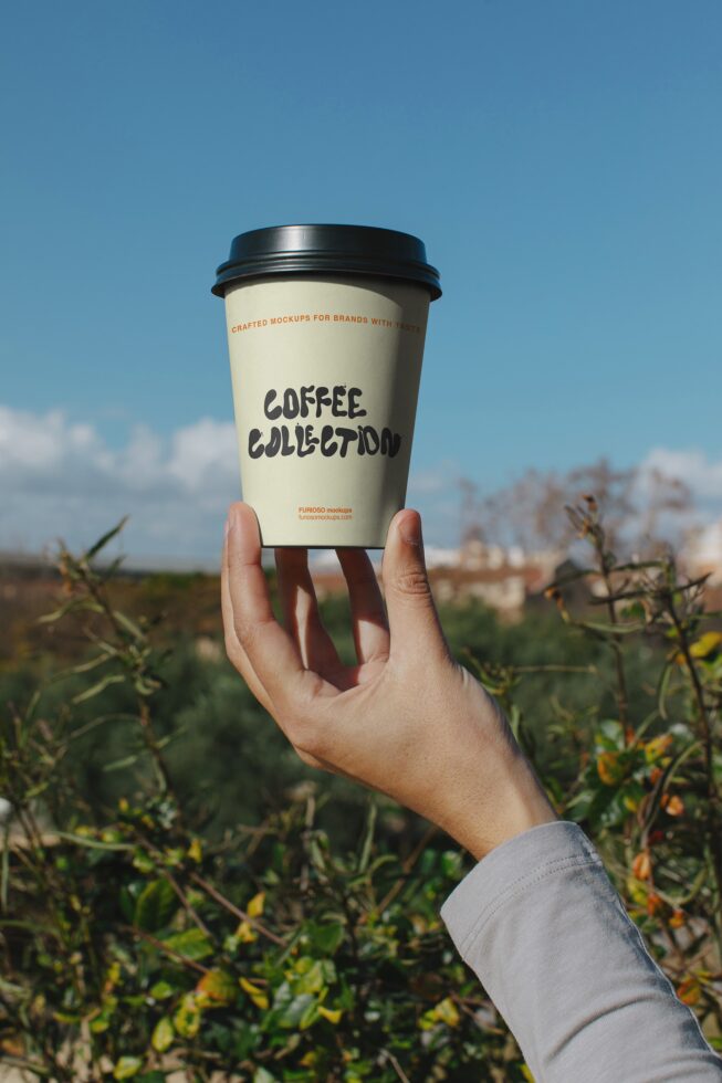 Hand holding cream coffee cup with black lid outdoors against blue sky