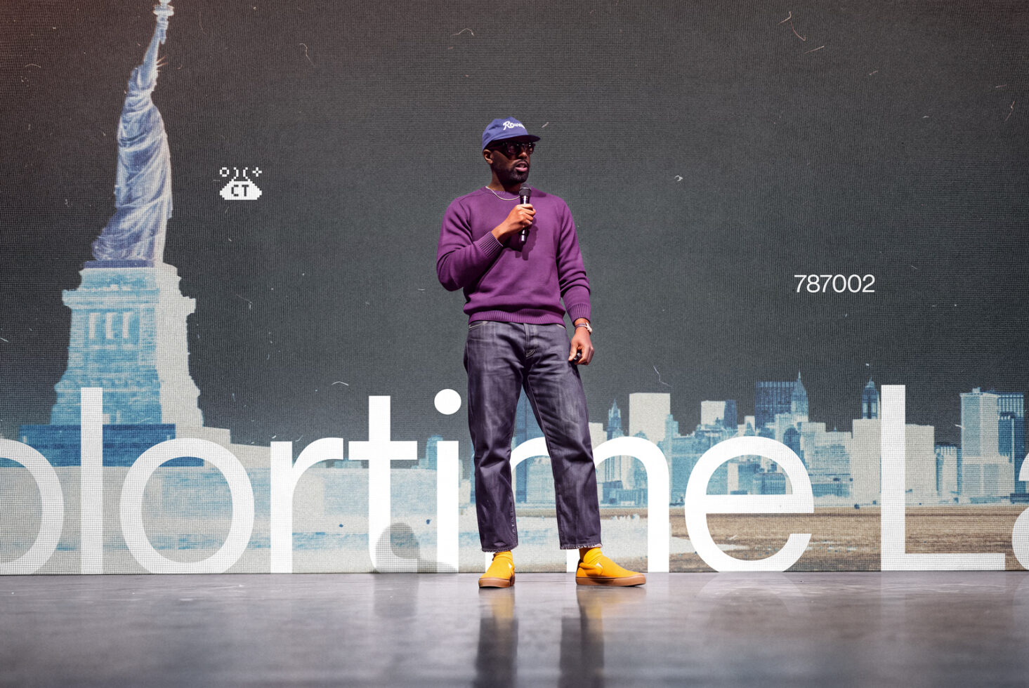 Man speaking on stage with NYC skyline backdrop, purple sweatshirt, yellow shoes