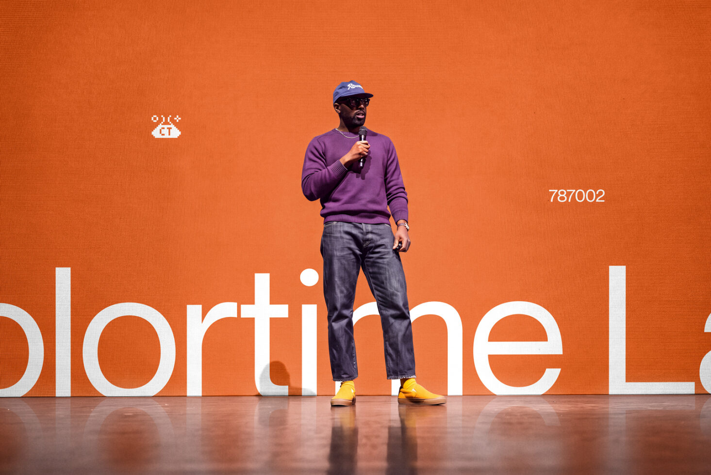 Guy on stage holding mic in purple crewneck, blue cap, yellow sneakers against bold orange backdrop