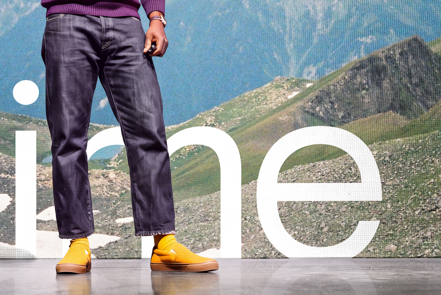 Person wearing dark jeans, purple ribbed sweater, yellow sneakers standing in front of mountain landscape with large white typography overlay
