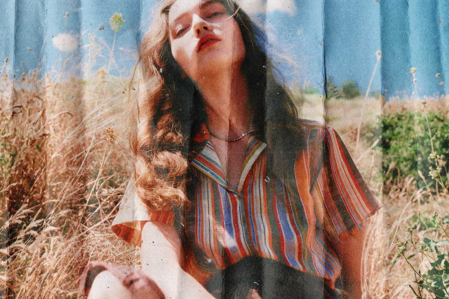 Woman in striped shirt sitting in dry grass with blue wall behind, vintage film photo aesthetic