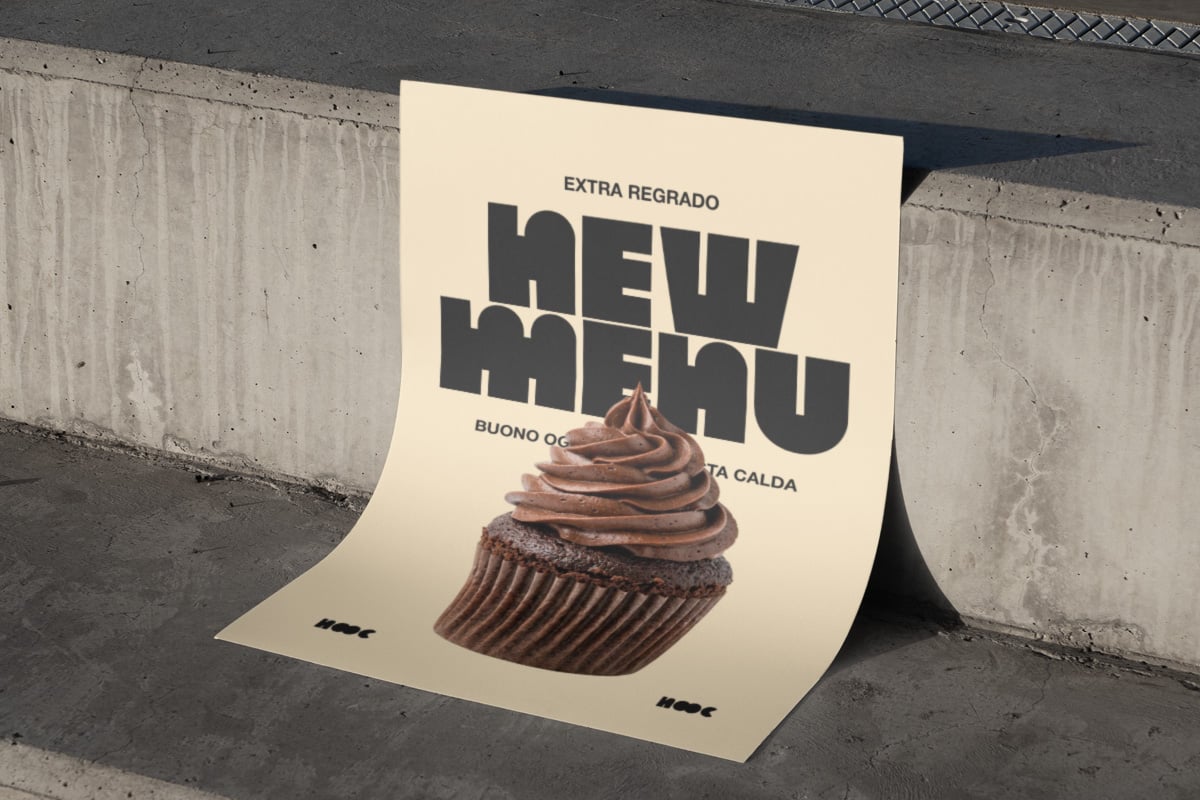 Bent cream poster with bold black typography leaning against concrete wall, chocolate cupcake photo centered below text