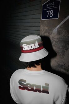 Person wearing white Seoul bucket hat with red stripe against grungy wall with street sign
