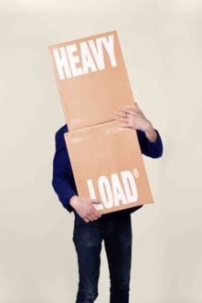 Person holding stacked cardboard boxes with "HEAVY LOAD" text, navy sleeve visible