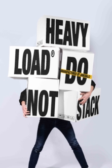 Person holding stacked cardboard boxes with bold black text reading "Heavy Load Do Not Stack"