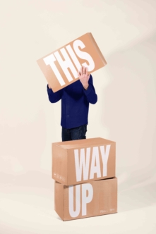 Person stacked on cardboard boxes holding sign reading "This Way Up"