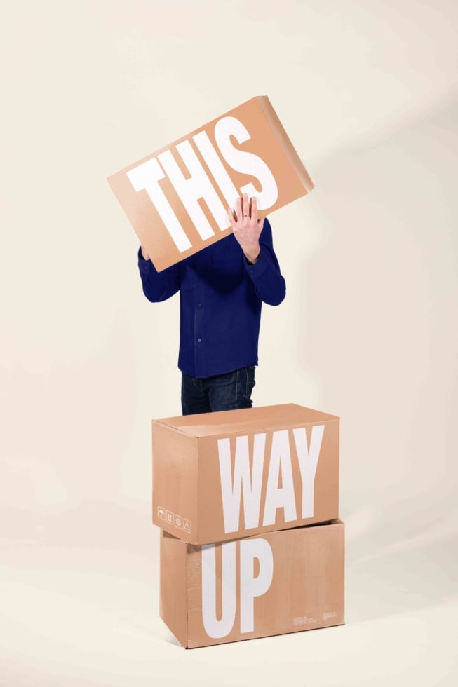 Person stacked on cardboard boxes holding sign reading "This Way Up"