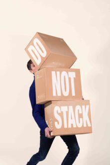 Guy struggling to carry three stacked cardboard boxes labeled "Do Not Stack"