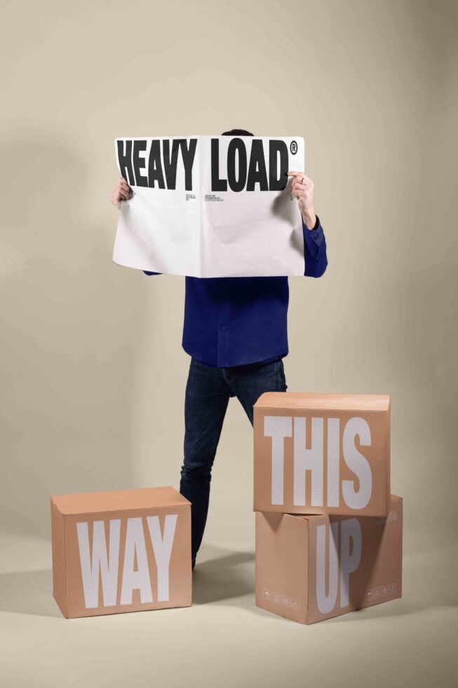 Person holding Heavy Load poster above stacked cardboard boxes saying Way Up This Up