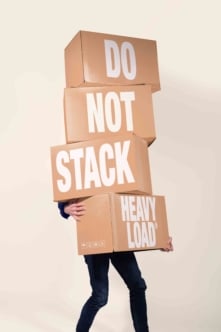 Person carrying precariously stacked cardboard boxes with "do not stack heavy load" text warning