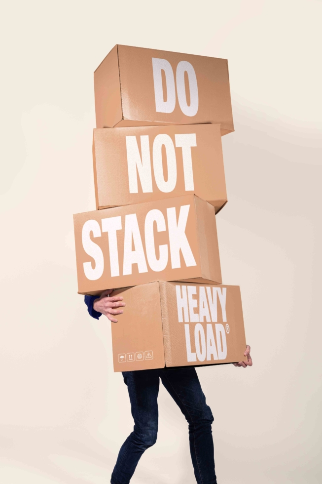 Person carrying precariously stacked cardboard boxes with "do not stack heavy load" text warning