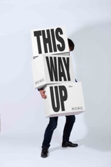 Person holding stacked cardboard boxes with bold black typography reading "THIS WAY UP"