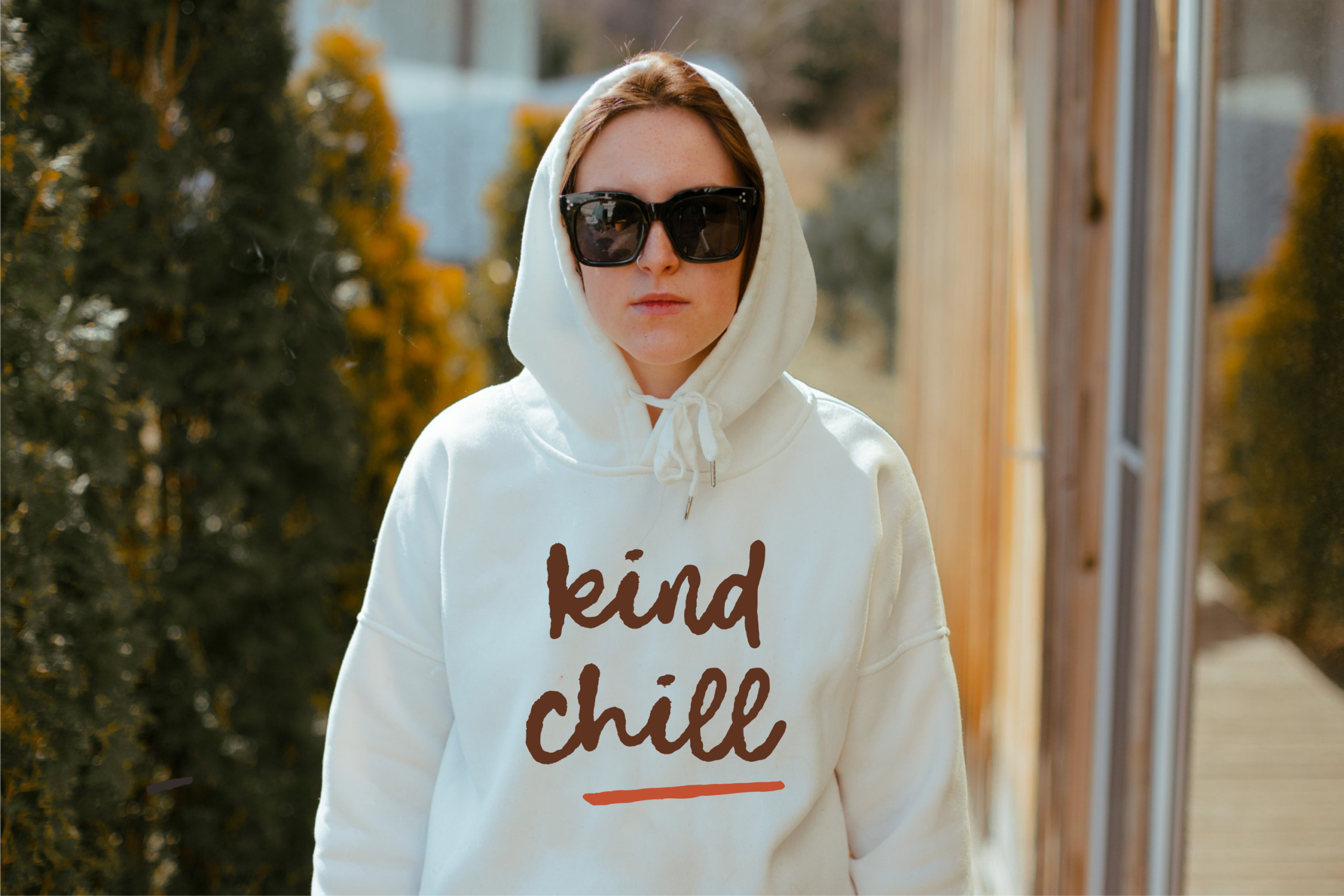Woman wearing cream hoodie with "kind chill" text design, black sunglasses, outdoor setting with blurred greenery