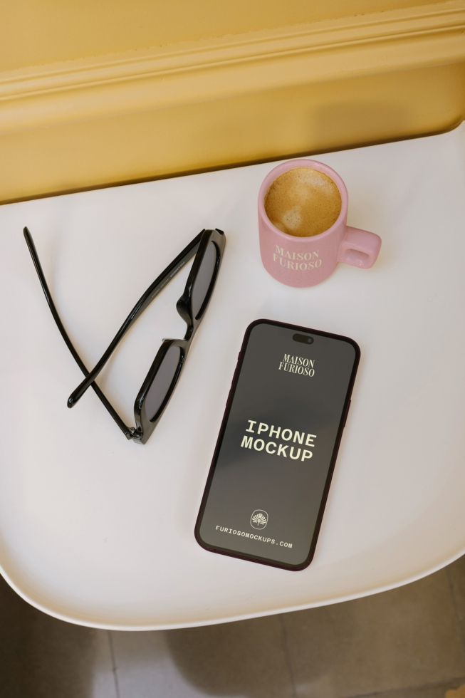 iPhone mockup styled with coffee cup, sunglasses, gold accents on white surface