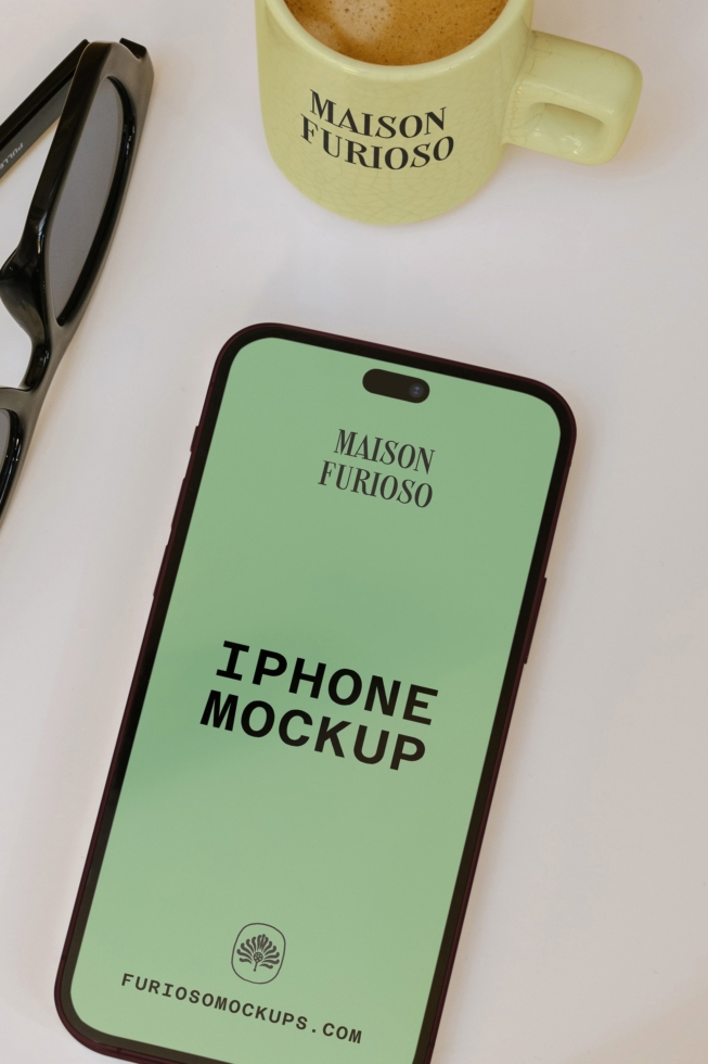 iPhone mockup with sage green screen showing Maison Furioso branding, styled with sunglasses plus coffee mug