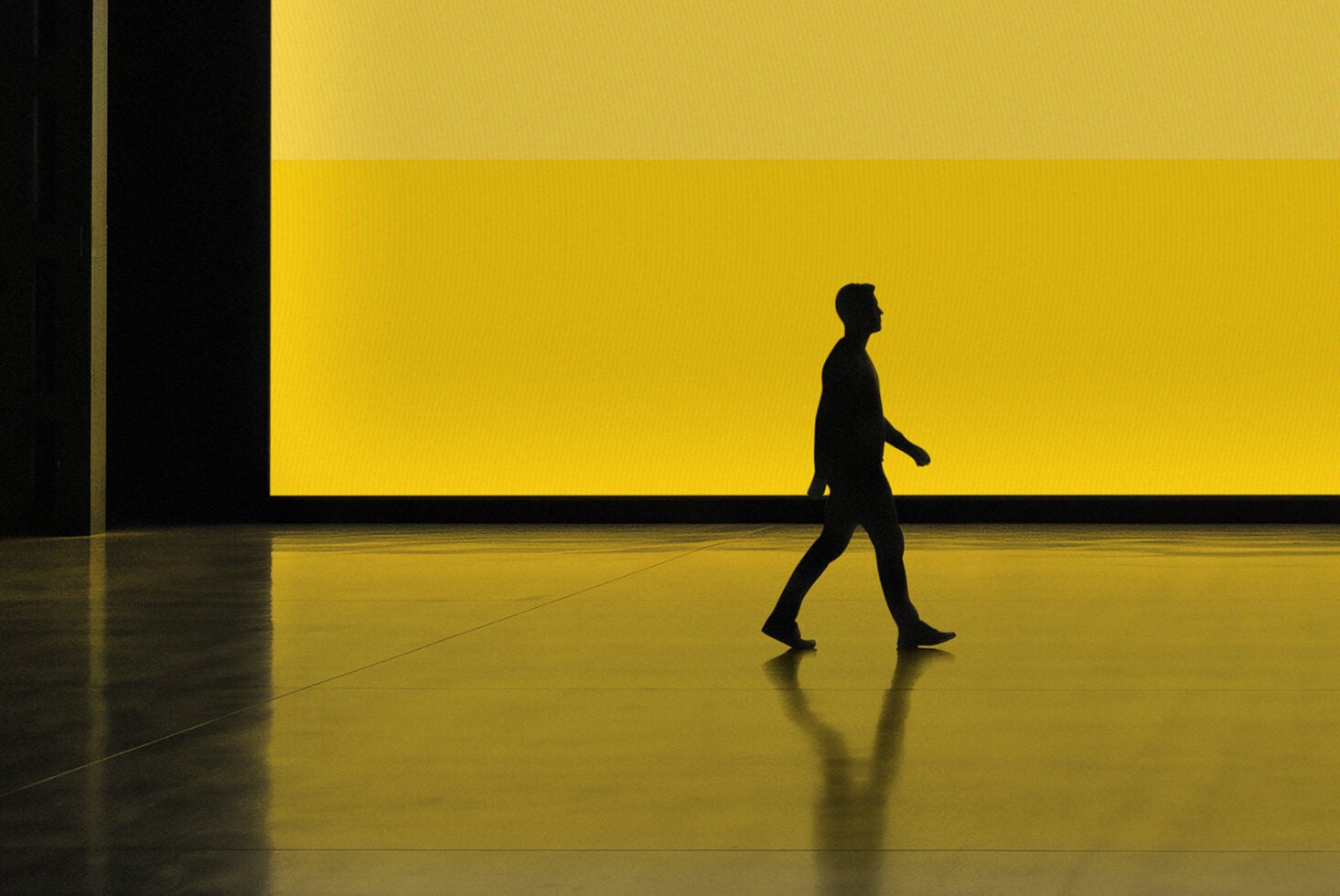 Silhouette of person walking across reflective floor with bold yellow wall behind them