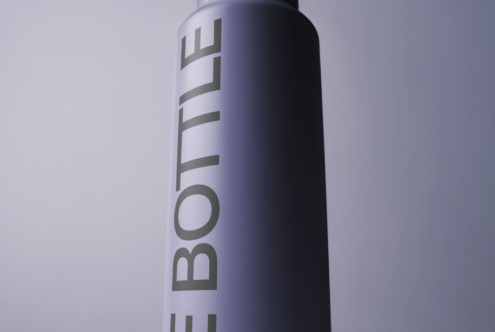 Tall spray bottle mockup with gradient fade from white to dark purple, vertical text wrapping around cylinder