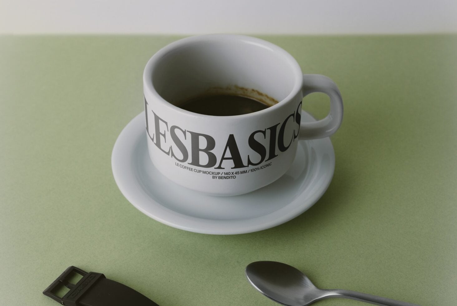 Ceramic coffee cup with saucer filled with espresso, labeled "LESBASICO", sitting on sage green background with spoon