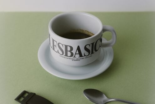 Ceramic coffee cup with saucer filled with espresso, labeled "LESBASICO", sitting on sage green background with spoon