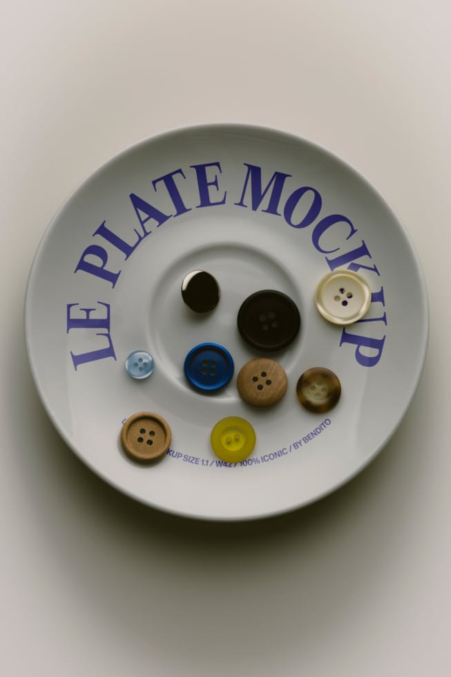 Overhead shot of ceramic plate mockup displaying assorted buttons in wood, plastic, black with blue text reading "LE PLATE MOCKUP"