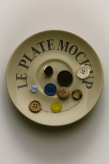 Overhead shot of cream ceramic plate with mixed buttons in various sizes, colors, wood finishes arranged inside curved text reading "Le Plate Mockup"