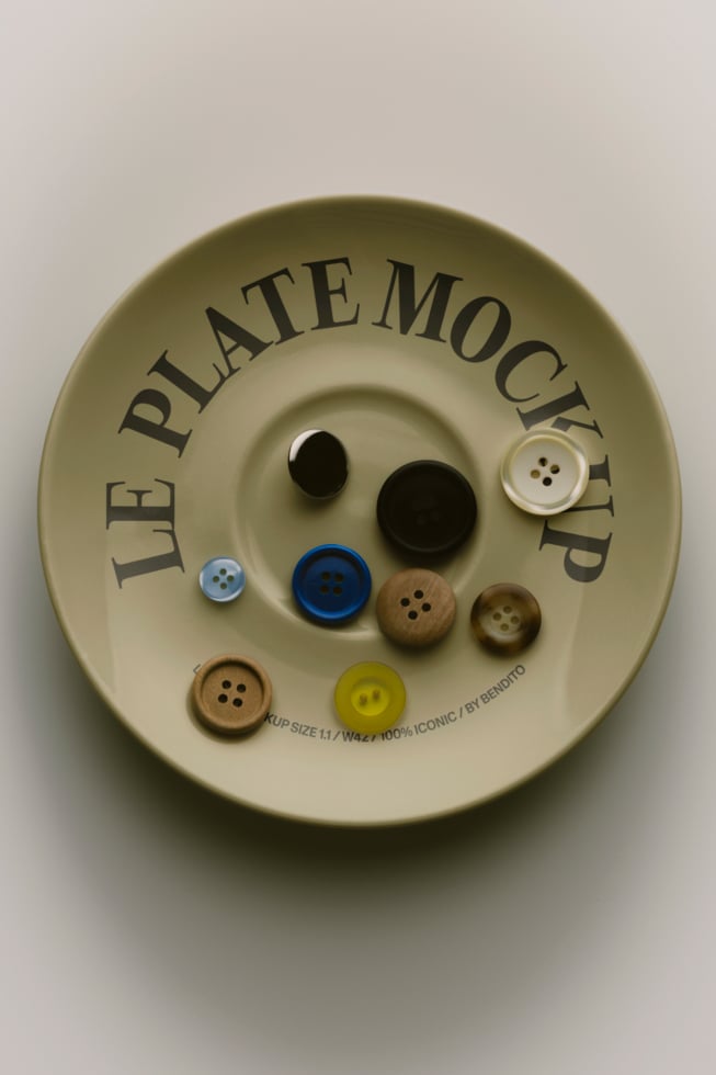 Overhead shot of cream ceramic plate with mixed buttons in various sizes, colors, wood finishes arranged inside curved text reading "Le Plate Mockup"