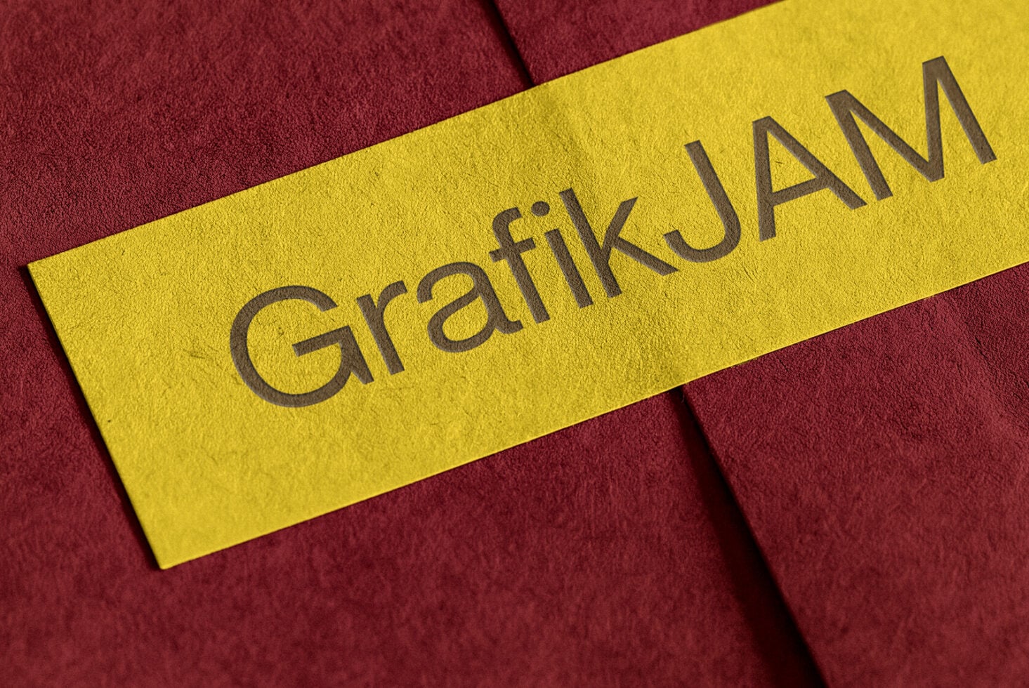Bright yellow paper strip with embossed gray text reading "Graffik Jam" laid diagonally on deep burgundy fabric background