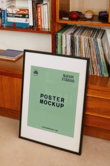 Black framed poster leaning against mid-century wood shelving with books, CDs, vinyl records displayed above
