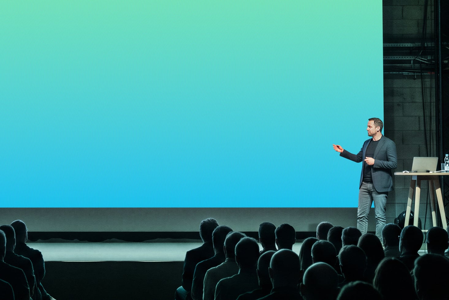 Guy presenting on stage with massive turquoise screen behind him, audience silhouettes in foreground