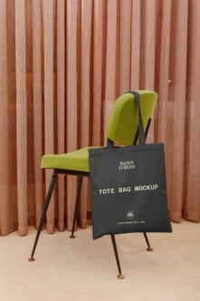 Black tote bag mockup draped over lime green mid-century chair with terracotta curtains