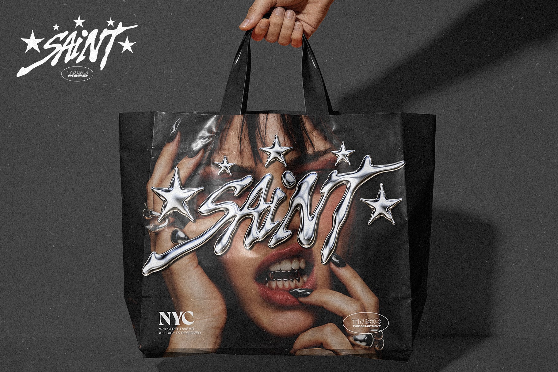 Hand holding black tote bag with edgy Saint branding, chrome stars, bold typography on dark moody background